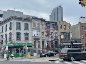 A city street with a row of diverse architectural buildings including a halal Chinese food restaurant, colorful murals on one building, and a deli. Pedestrians are walking on the sidewalk, and a few vehicles are visible on the street under a cloudy sky.