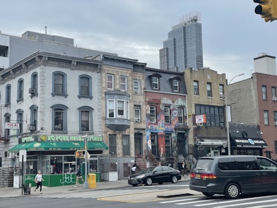 A city street with a row of diverse architectural buildings including a halal Chinese food restaurant, colorful murals on one building, and a deli. Pedestrians are walking on the sidewalk, and a few vehicles are visible on the street under a cloudy sky.