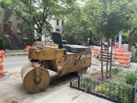 A construction site on a city street features a large yellow steamroller parked next to a tree and some orange traffic barrels. Nearby, there are stacks of construction materials and barriers. Residential buildings and greenery surround the area, indicating an urban setting.