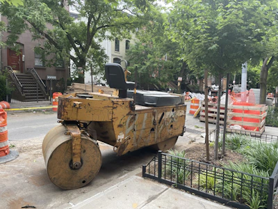 A construction site on a city street features a large yellow steamroller parked next to a tree and some orange traffic barrels. Nearby, there are stacks of construction materials and barriers. Residential buildings and greenery surround the area, indicating an urban setting.