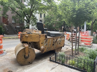 A construction site on a city street features a large yellow steamroller parked next to a tree and some orange traffic barrels. Nearby, there are stacks of construction materials and barriers. Residential buildings and greenery surround the area, indicating an urban setting.