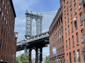 A steel suspension bridge is framed between two red brick buildings. The bridge features large, intricate metalwork with suspension cables extending upwards into a partly cloudy blue sky. Below, there is a street sign and some greenery.