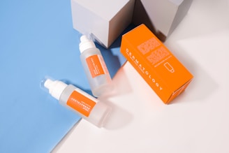 Two translucent bottles with orange labels and white caps are placed on a light blue and white surface. Next to the bottles is an orange rectangular box with white text that reads 'DERMATOLOGY'. The setting includes geometric shadows from the light source.