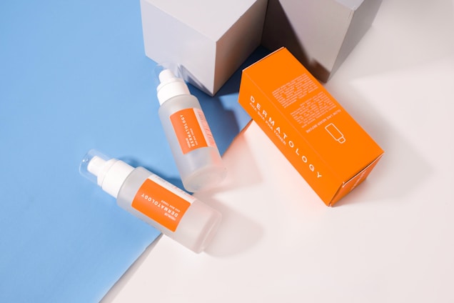 Two translucent bottles with orange labels and white caps are placed on a light blue and white surface. Next to the bottles is an orange rectangular box with white text that reads 'DERMATOLOGY'. The setting includes geometric shadows from the light source.