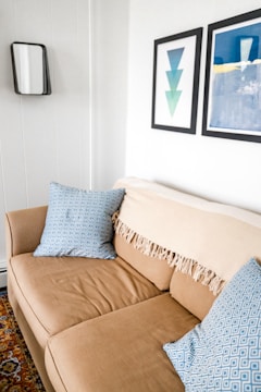 A cozy beige sofa is adorned with two blue patterned cushions. A tasseled beige throw is draped over the backrest. The white walls are decorated with minimalist art prints; one features overlapping triangles, and the other displays abstract blue and white forms. A small, rectangular mirror with a black frame hangs nearby, and part of a colorful patterned rug is visible on the floor.