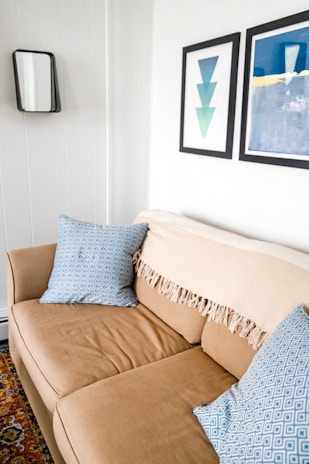 A cozy beige sofa is adorned with two blue patterned cushions. A tasseled beige throw is draped over the backrest. The white walls are decorated with minimalist art prints; one features overlapping triangles, and the other displays abstract blue and white forms. A small, rectangular mirror with a black frame hangs nearby, and part of a colorful patterned rug is visible on the floor.