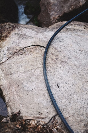 A black hose lies on a rough, large rock surface. The rock has a crack running through it and is surrounded by other rocks. There is a small amount of water and debris around the base of the rocks.