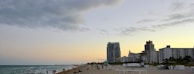A sandy beach with people enjoying leisure activities. The beach features several groups of people scattered along the shoreline. In the background, modern high-rise buildings line the coast, reflecting an urban beach setting. The sky is partially cloudy with a hint of sunset, creating a serene atmosphere.