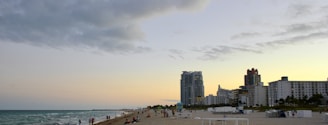 A sandy beach with people enjoying leisure activities. The beach features several groups of people scattered along the shoreline. In the background, modern high-rise buildings line the coast, reflecting an urban beach setting. The sky is partially cloudy with a hint of sunset, creating a serene atmosphere.
