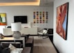 A modern living room with contemporary art on the walls, including a large painting of a face blowing bubbles. The room features a sleek, minimalist design with a black and white color scheme. A spherical, metallic table lamp and a geometric-patterned rug add decorative elements. There is also a wall-mounted flat-screen TV and an array of colorful square tiles as artwork.