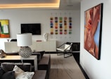 A modern living room with contemporary art on the walls, including a large painting of a face blowing bubbles. The room features a sleek, minimalist design with a black and white color scheme. A spherical, metallic table lamp and a geometric-patterned rug add decorative elements. There is also a wall-mounted flat-screen TV and an array of colorful square tiles as artwork.