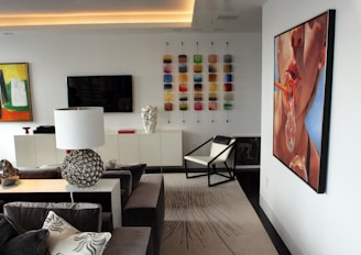 A modern living room with contemporary art on the walls, including a large painting of a face blowing bubbles. The room features a sleek, minimalist design with a black and white color scheme. A spherical, metallic table lamp and a geometric-patterned rug add decorative elements. There is also a wall-mounted flat-screen TV and an array of colorful square tiles as artwork.