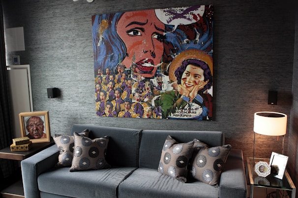 A stylish living room features a prominent, colorful pop-art style painting hanging above a modern grey sofa adorned with patterned cushions. To the left is a small side table with decorative items including a framed portrait, and on the right is a glass side table with a round lamp and framed photograph. The overall decor is contemporary with dark tones and artistic flair.