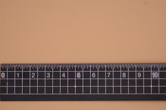 A black ruler with white markings is placed on a flat light-colored surface. It features a grid pattern with both numerical and fractional increments.