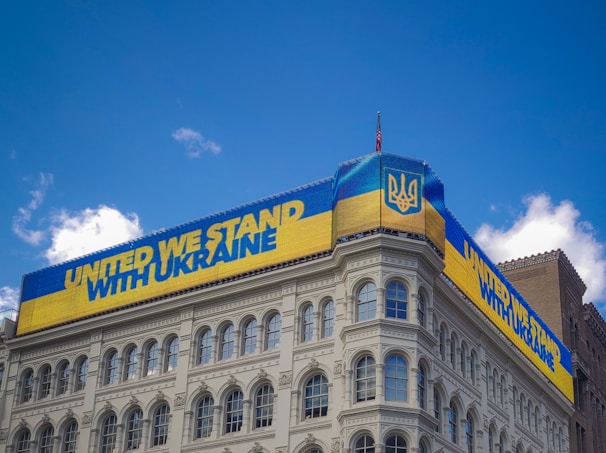 A building displays a large digital banner with a message of solidarity for Ukraine. The banner is in the colors of the Ukrainian flag, featuring blue and yellow stripes and the national emblem. The sky is clear with a few white clouds.