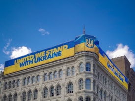 A building displays a large digital banner with a message of solidarity for Ukraine. The banner is in the colors of the Ukrainian flag, featuring blue and yellow stripes and the national emblem. The sky is clear with a few white clouds.