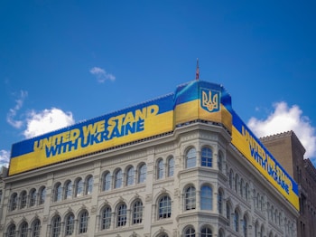 A building displays a large digital banner with a message of solidarity for Ukraine. The banner is in the colors of the Ukrainian flag, featuring blue and yellow stripes and the national emblem. The sky is clear with a few white clouds.