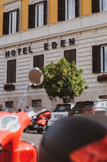 A street view featuring a section of a building with the words 'Hotel Eden' displayed prominently. The facade is cream-colored with contrasting dark window shutters. In front of the building, there is a vibrant orange tree and a row of parked vehicles, including a red scooter in the foreground.