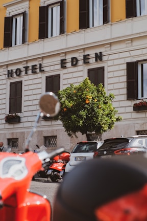 A street view featuring a section of a building with the words 'Hotel Eden' displayed prominently. The facade is cream-colored with contrasting dark window shutters. In front of the building, there is a vibrant orange tree and a row of parked vehicles, including a red scooter in the foreground.