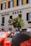 A street view featuring a section of a building with the words 'Hotel Eden' displayed prominently. The facade is cream-colored with contrasting dark window shutters. In front of the building, there is a vibrant orange tree and a row of parked vehicles, including a red scooter in the foreground.