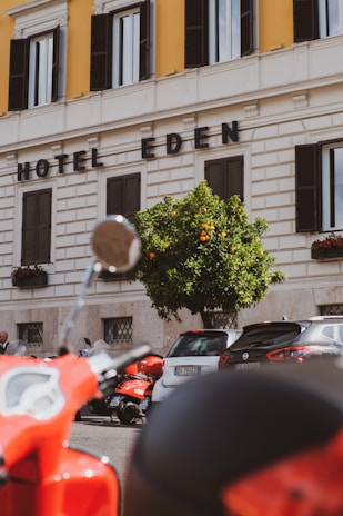 A street view featuring a section of a building with the words 'Hotel Eden' displayed prominently. The facade is cream-colored with contrasting dark window shutters. In front of the building, there is a vibrant orange tree and a row of parked vehicles, including a red scooter in the foreground.