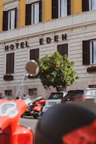 A street view featuring a section of a building with the words 'Hotel Eden' displayed prominently. The facade is cream-colored with contrasting dark window shutters. In front of the building, there is a vibrant orange tree and a row of parked vehicles, including a red scooter in the foreground.