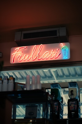 A neon sign displaying the word 'Frullati' is mounted above a storefront. The sign also features a neon graphic of a drink in a cup with a straw. Below the sign, there are slush machines, cups, and other beverage-related items.