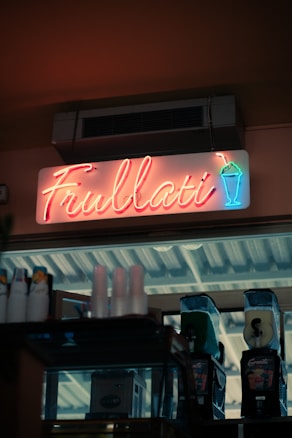 A neon sign displaying the word 'Frullati' is mounted above a storefront. The sign also features a neon graphic of a drink in a cup with a straw. Below the sign, there are slush machines, cups, and other beverage-related items.