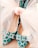 A pair of intricately patterned high-heeled shoes adorned with a large, delicate, pale pink ribbon tied in a bow. The shoes display a distinctive geometric design in shades of teal and black, creating a striking visual contrast.