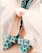 A pair of intricately patterned high-heeled shoes adorned with a large, delicate, pale pink ribbon tied in a bow. The shoes display a distinctive geometric design in shades of teal and black, creating a striking visual contrast.
