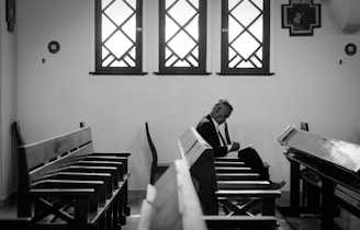A black and white image of a man sitting alone on a wooden pew inside a church or chapel. He appears to be deep in thought or prayer, possibly writing or reading from a notebook. The lighting is soft, coming through three large windows with decorative grilles behind him.