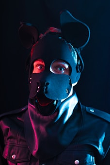 A person is wearing a black leather costume with canine-like features, including a mask with ears and facial details resembling a dog's snout. The lighting is dramatic with blue and pink tones, highlighting the texture of the leather material.