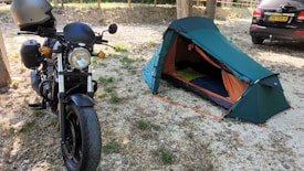A parked motorcycle with a helmet on the handlebar is adjacent to a small, pitched camping tent. The setting is outdoors on a gravel and grass area, with a car visible in the background to the right.