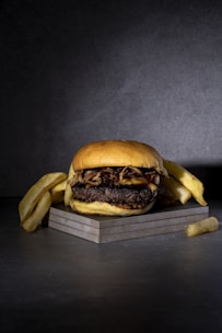 A hamburger with a thick beef patty, cheese, and grilled onions is placed on a square slate surface. It is accompanied by several thick-cut French fries arranged around it. The background is dark gray, adding a dramatic contrast to the food.