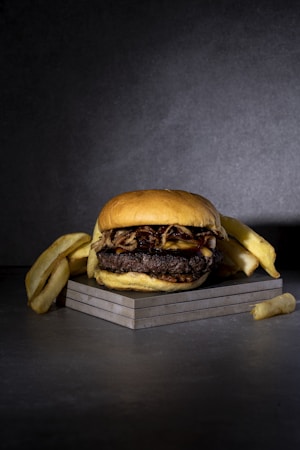 A hamburger with a thick beef patty, cheese, and grilled onions is placed on a square slate surface. It is accompanied by several thick-cut French fries arranged around it. The background is dark gray, adding a dramatic contrast to the food.