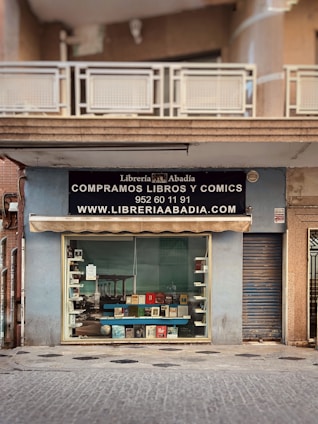 A storefront with a sign for 'Librería Abadía' offering books and comics for sale. The facade is light blue, featuring a large window display showcasing various books. Above the window, there is an awning and a black sign with white text including a phone number and website. The entrance is adjacent to a closed metal shutter.