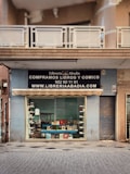 A storefront with a sign for 'Librería Abadía' offering books and comics for sale. The facade is light blue, featuring a large window display showcasing various books. Above the window, there is an awning and a black sign with white text including a phone number and website. The entrance is adjacent to a closed metal shutter.