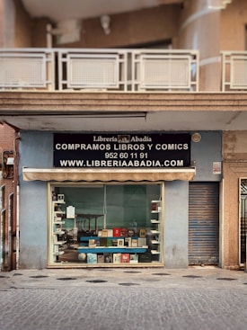 A storefront with a sign for 'Librer&iacute;a Abad&iacute;a' offering books and comics for sale. The facade is light blue, featuring a large window display showcasing various books. Above the window, there is an awning and a black sign with white text including a phone number and website. The entrance is adjacent to a closed metal shutter.