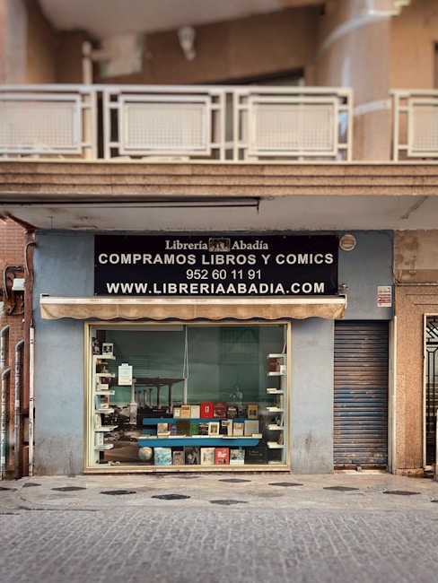 A storefront with a sign for 'Librería Abadía' offering books and comics for sale. The facade is light blue, featuring a large window display showcasing various books. Above the window, there is an awning and a black sign with white text including a phone number and website. The entrance is adjacent to a closed metal shutter.