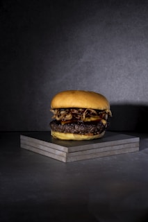 A single hamburger is placed on a stack of gray marble-like tiles against a dark gray background. The burger features a soft, golden-brown bun, a thick beef patty, melted cheese, and crispy fried onions.