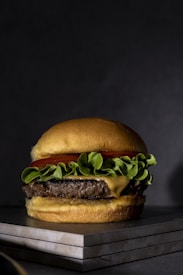 A juicy hamburger rests on a stack of tiles, featuring a perfectly grilled beef patty topped with a slice of melted cheese, fresh green lettuce, and a slice of red tomato. The burger is encased in a golden-brown bun, all against a dark, moody background.