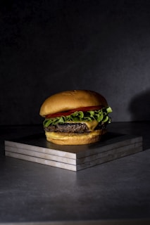 A hamburger with melted cheese, leafy green lettuce, and a slice of tomato stacked between a soft bun. It rests on a stack of three square tiles against a dark gray background, with moody lighting emphasizing the textures and colors of the ingredients.