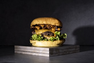 A juicy cheeseburger is stacked high with crisp lettuce, melted cheese, and a substantial patty between two soft buns. The background is simple and dark, making the burger the focal point, and it sits on a textured surface, possibly stone or slate, enhancing its presentation.