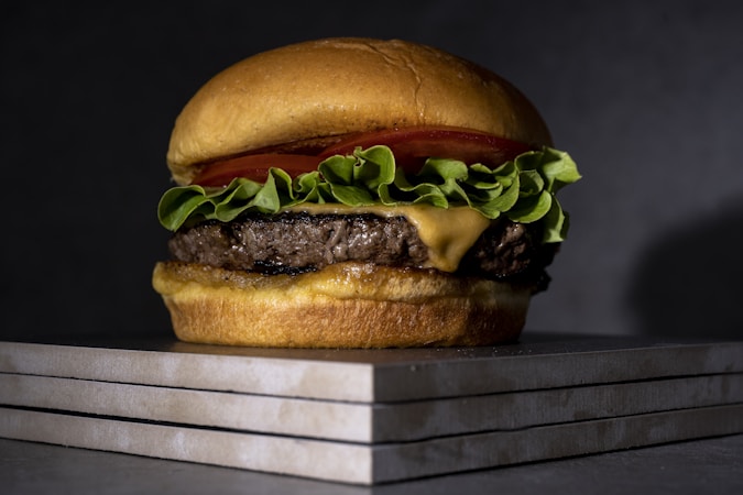 A close-up of a juicy hamburger with a beef patty, melted cheese, fresh lettuce, and a slice of tomato, all sandwiched between a golden-brown bun. The burger is resting on a stack of stone tiles, giving it an elegant presentation.