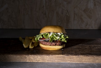 A hamburger with layers of lettuce, tomato, and mayonnaise sits on a rustic wooden board. To its left, a few golden-brown fries are visible against a dark, textured background.
