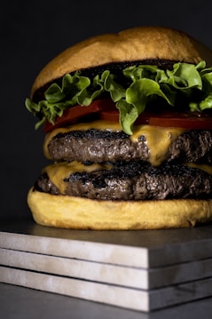 A close-up shot of a delicious double cheeseburger featuring two juicy beef patties topped with melted cheese. Crisp green lettuce and fresh red tomato slices are neatly layered under a soft toasted bun. The burger rests on a stack of flat, gray stone tiles, adding a modern and rustic touch to the presentation.