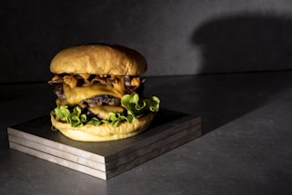A juicy cheeseburger featuring layers of melted cheese, crispy bacon, and fresh lettuce is placed on a wooden block. The lighting casts a dramatic shadow against a dark, textured background, highlighting the burger's textures.