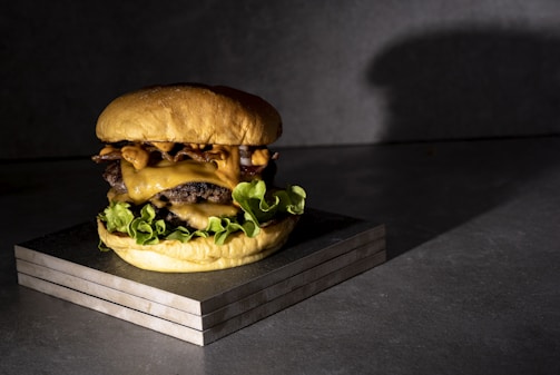 A juicy cheeseburger featuring layers of melted cheese, crispy bacon, and fresh lettuce is placed on a wooden block. The lighting casts a dramatic shadow against a dark, textured background, highlighting the burger's textures.