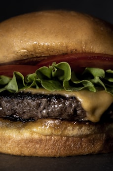 A close-up of a juicy cheeseburger with melted cheese, fresh lettuce, and tomato on a toasted bun, styled on a dark background with yellow and red accents.