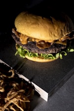 Close-up of a juicy classic burger on a black slate plate with fries on the side under warm lighting.